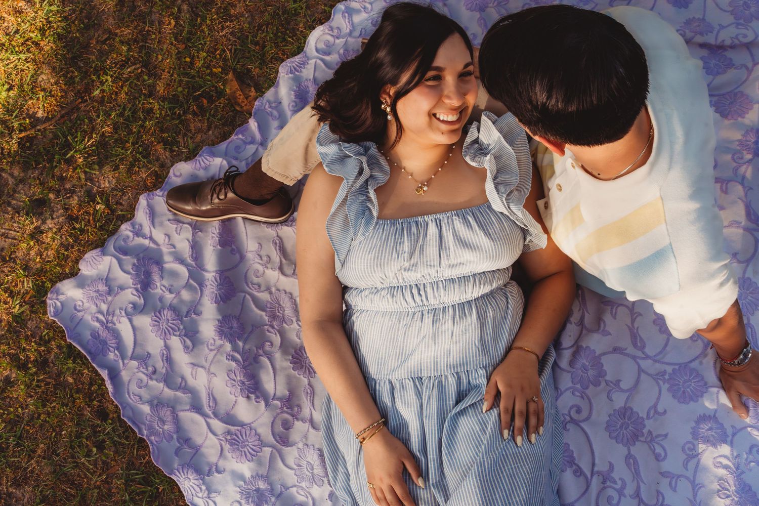 Couple lying on a purple blanket outdoors, smiling and sharing a romantic moment in warm sunlight.