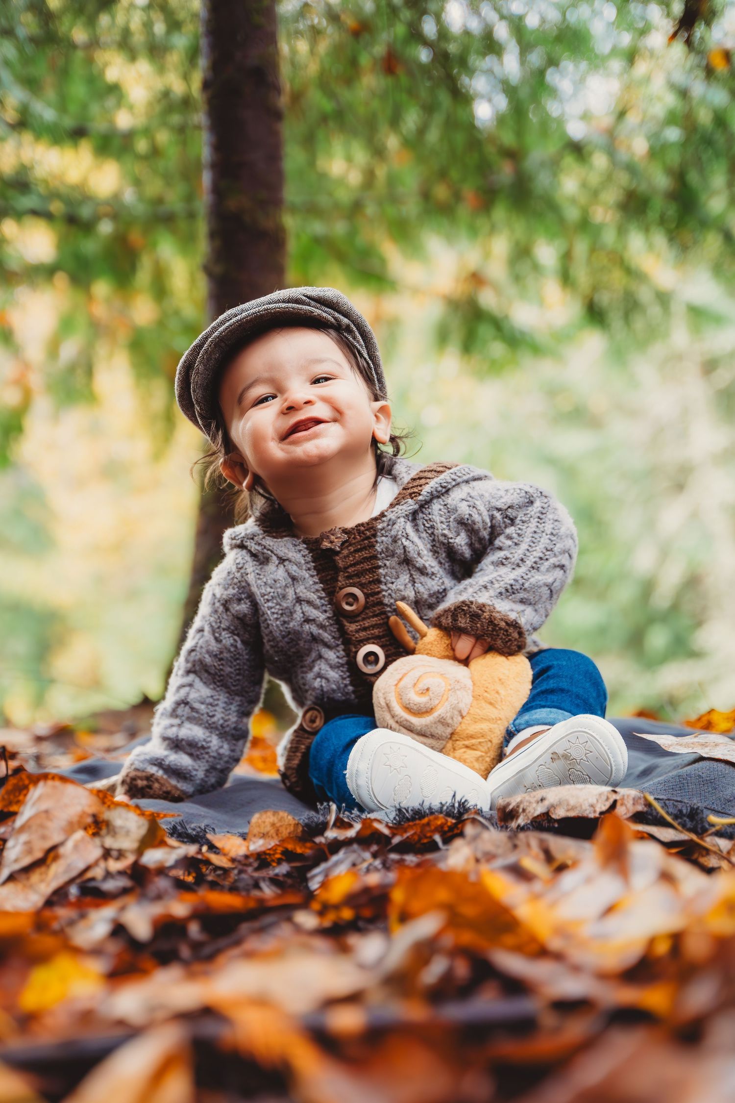Happy toddler in gray knit sweater and flat cap sits among autumn leaves in forest holding a stuffed animal toy.