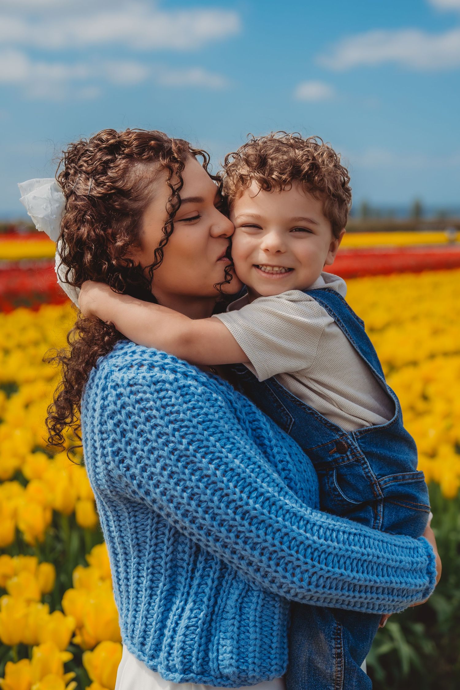 Mother kissing her smiling curly-haired son in a vibrant yellow and red tulip field under a blue sky.