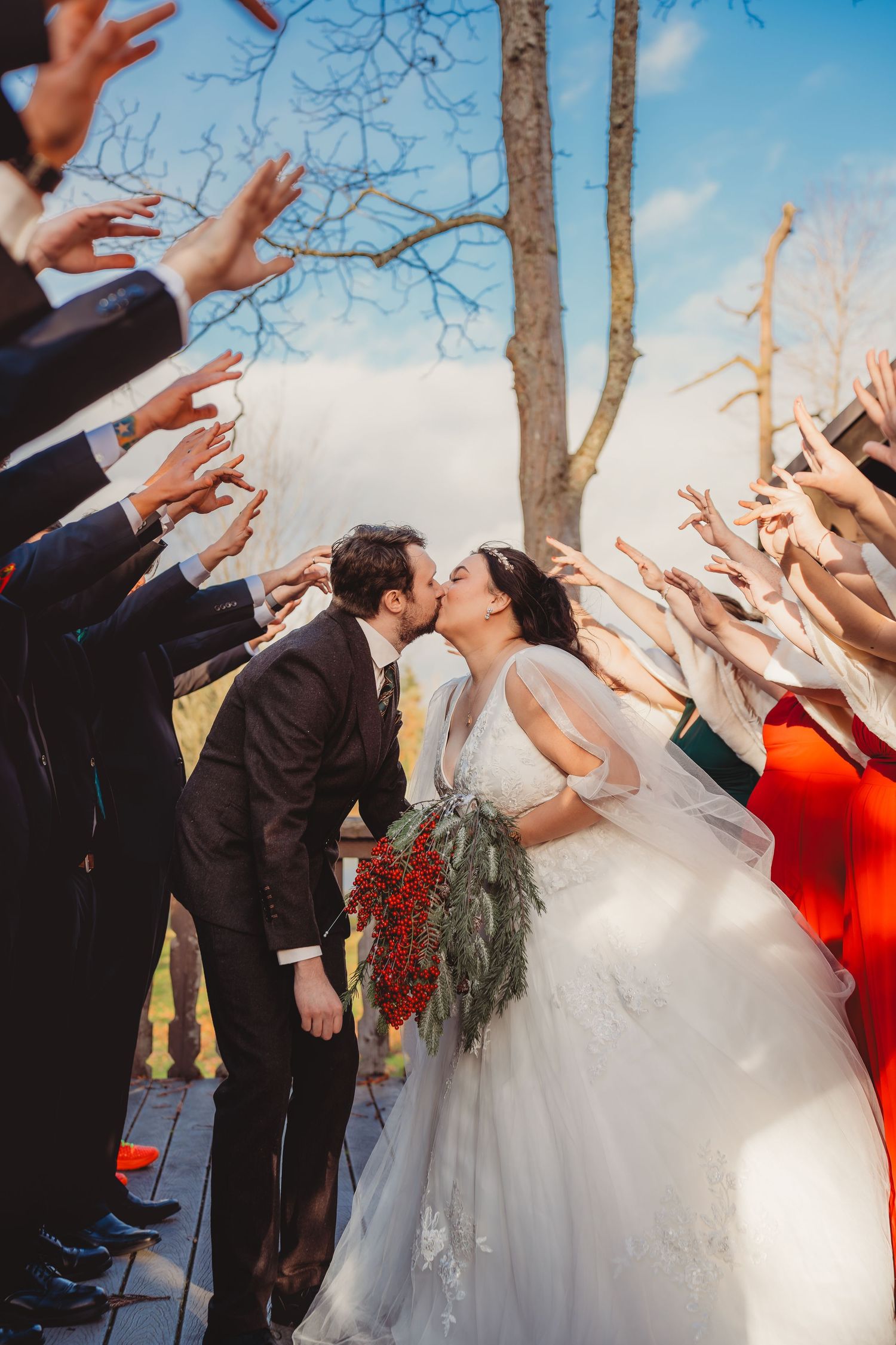 Bride and groom share a kiss while wedding guests raise their hands in celebration outdoors near a bare tree.