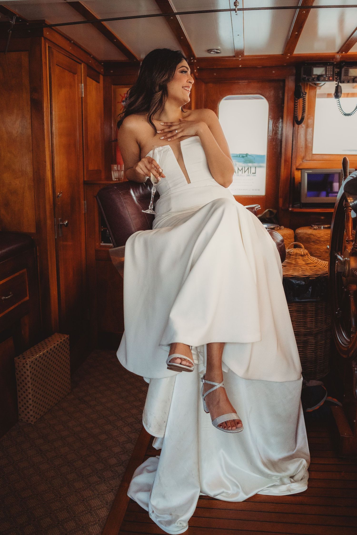 Bride in a white strapless gown sitting inside a yacht cabin, smiling and holding a glass.