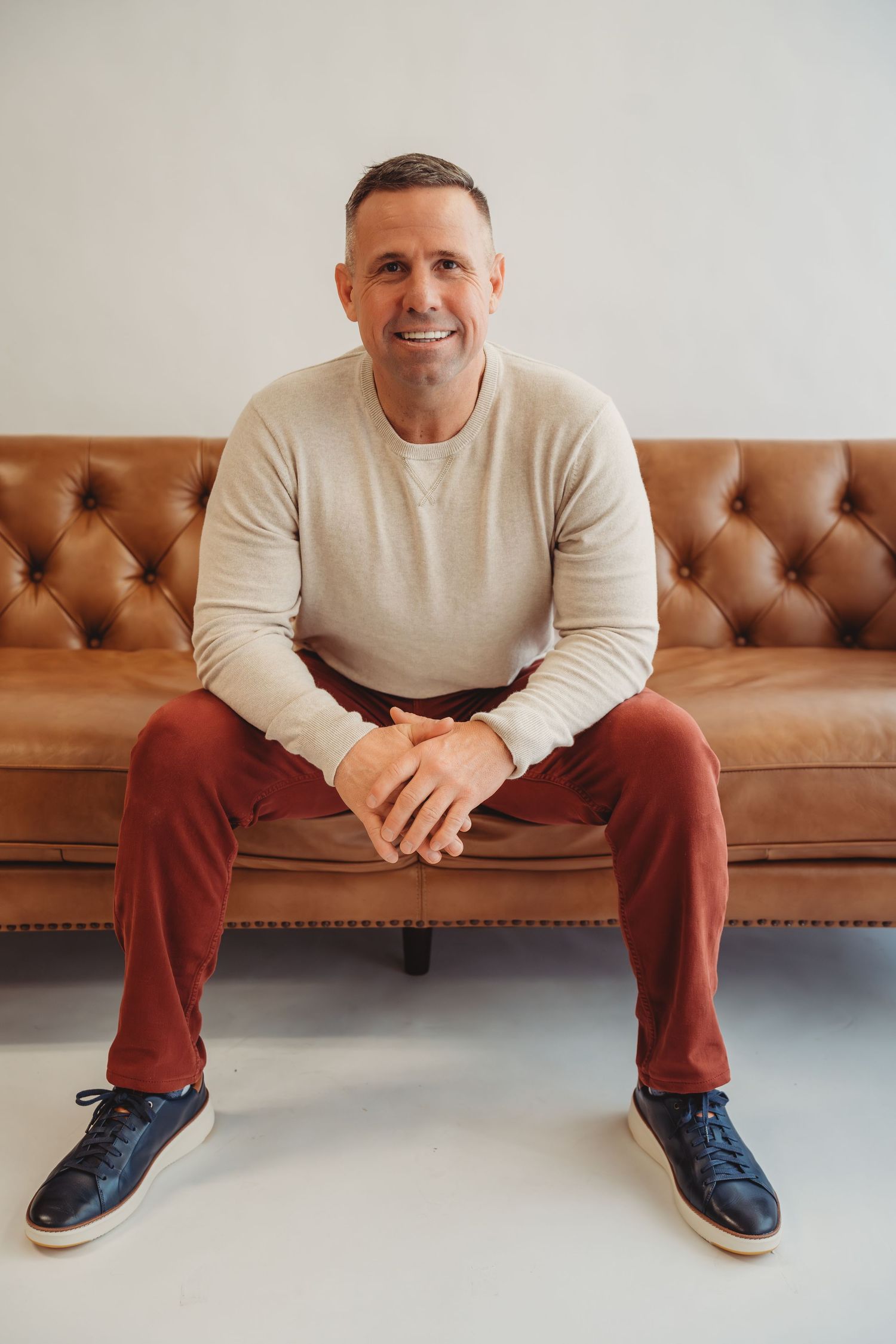 Smiling middle-aged man in beige sweater and red pants sitting on a tufted brown leather sofa.