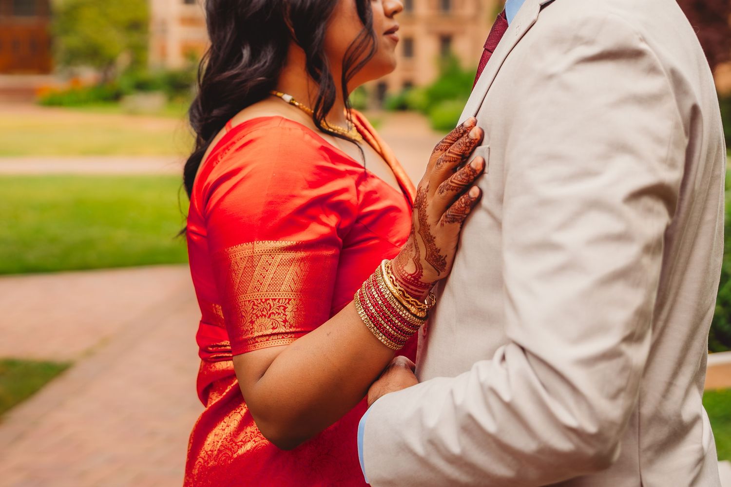 Indian bride in red and gold saree with bangles embraces groom in white suit during outdoor wedding ceremony.