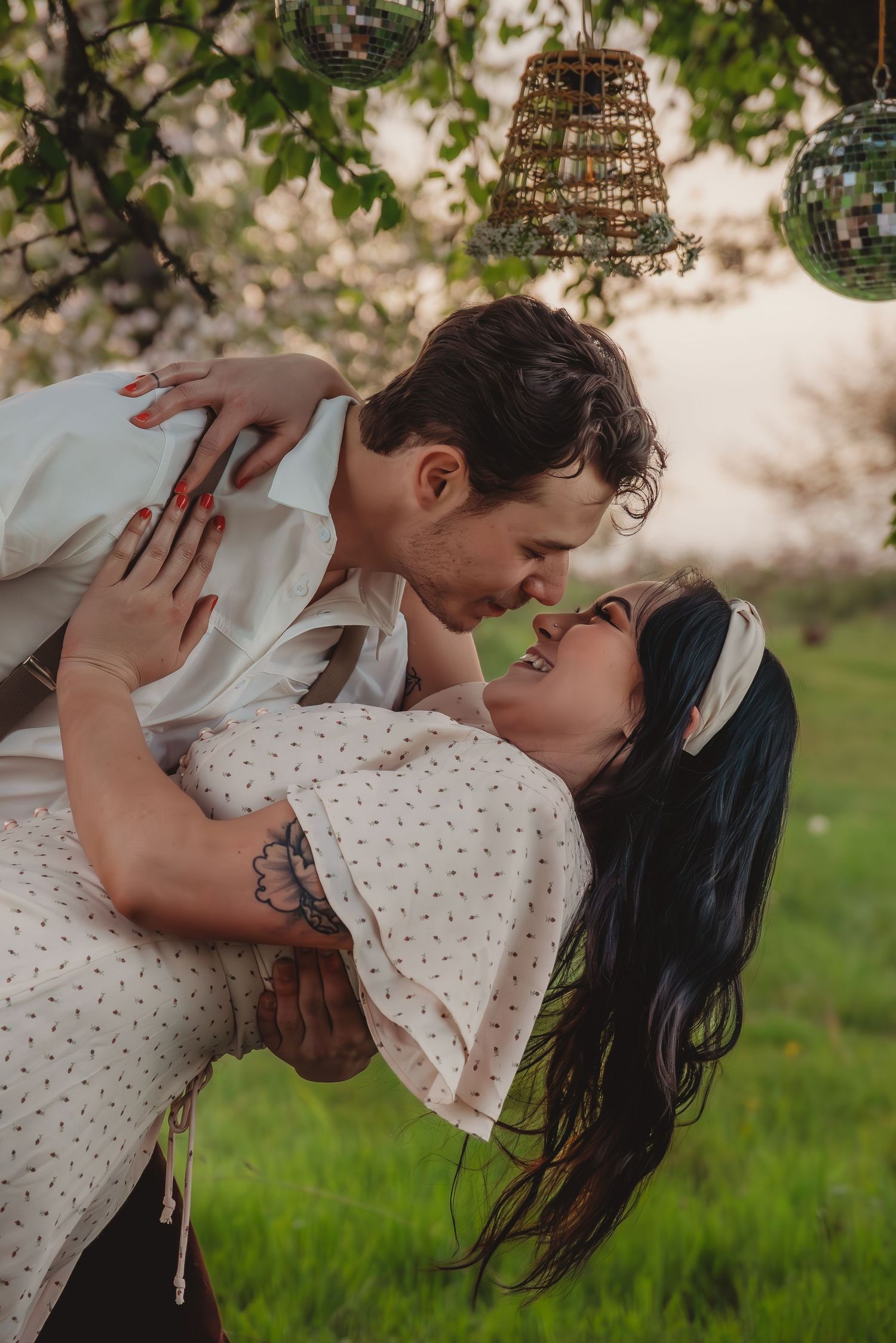 Man dipping woman in a romantic embrace under hanging disco balls in a blooming garden.