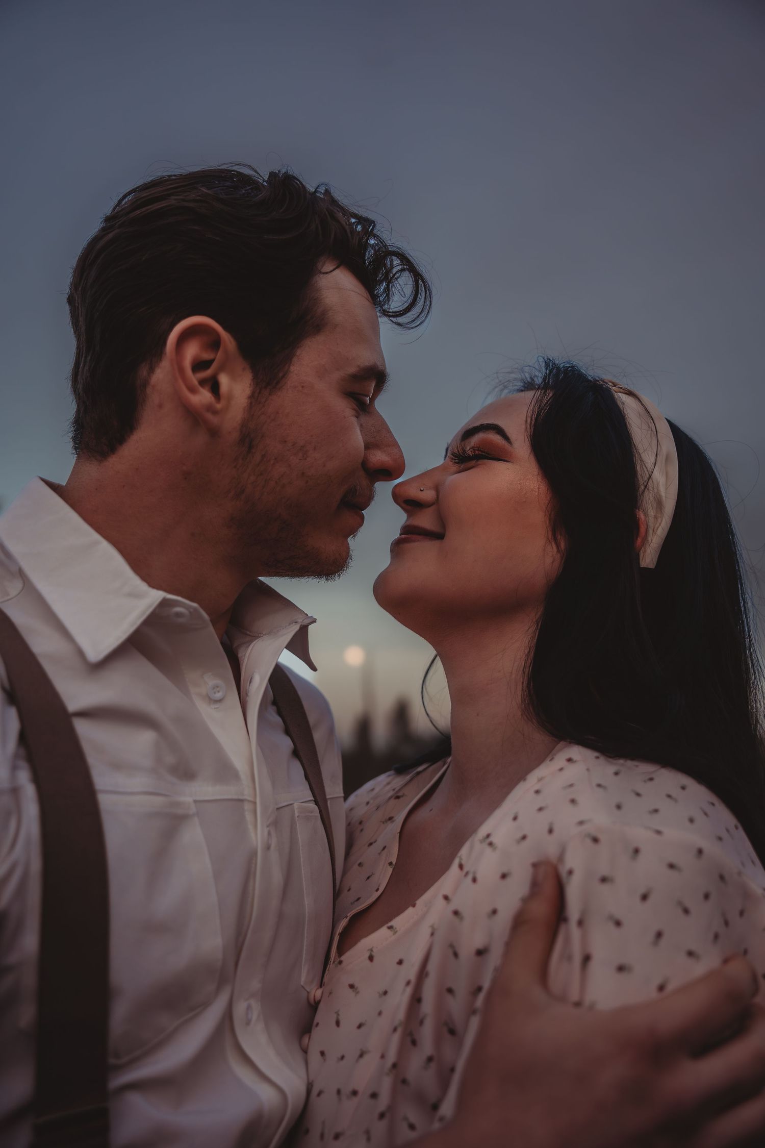 Couple sharing a tender almost-kiss moment outdoors at dusk with moonlit sky behind them.