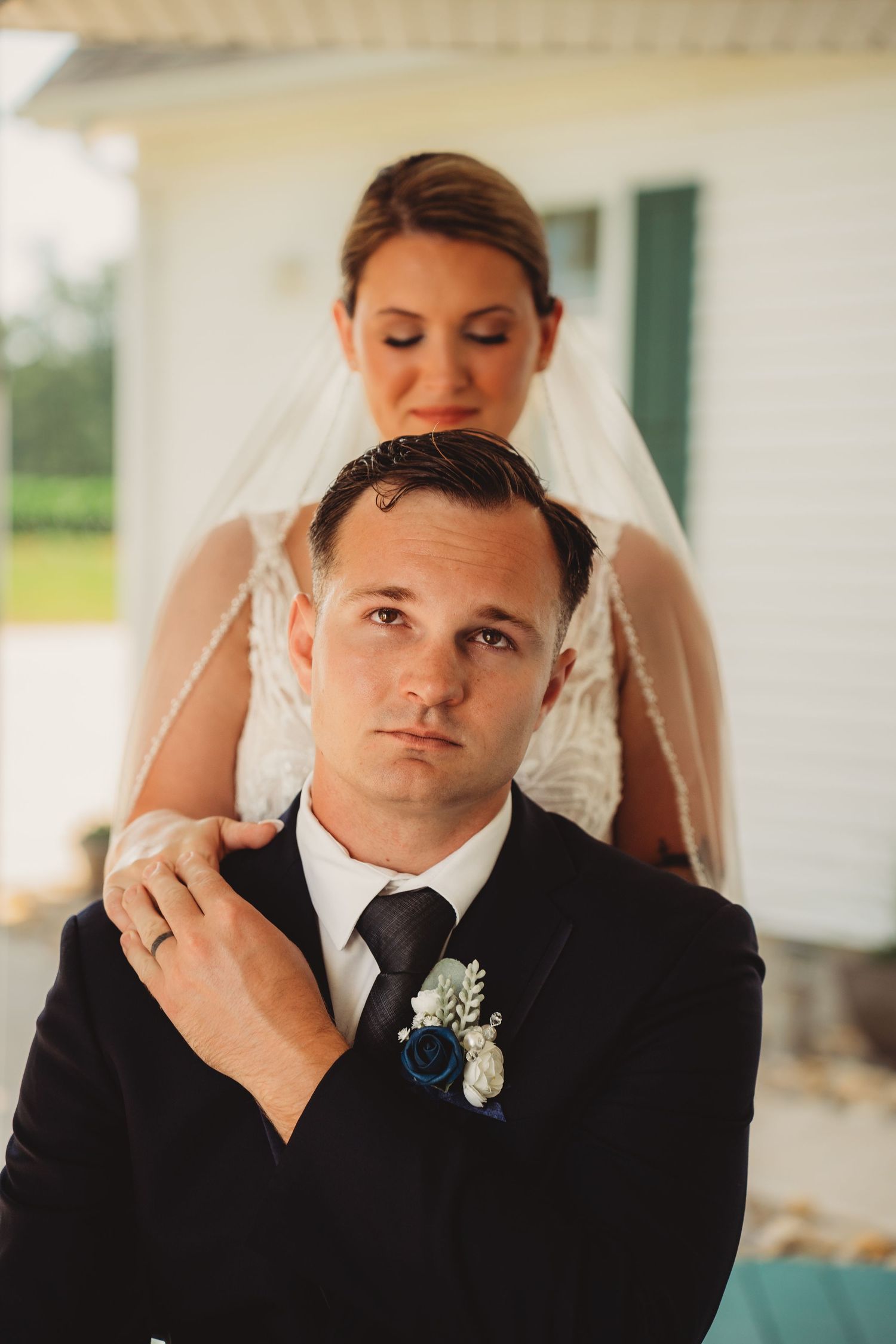 Bride in lace veil gently places hand on seated grooms shoulder in intimate portrait outside white farmhouse. Uptown Indiana