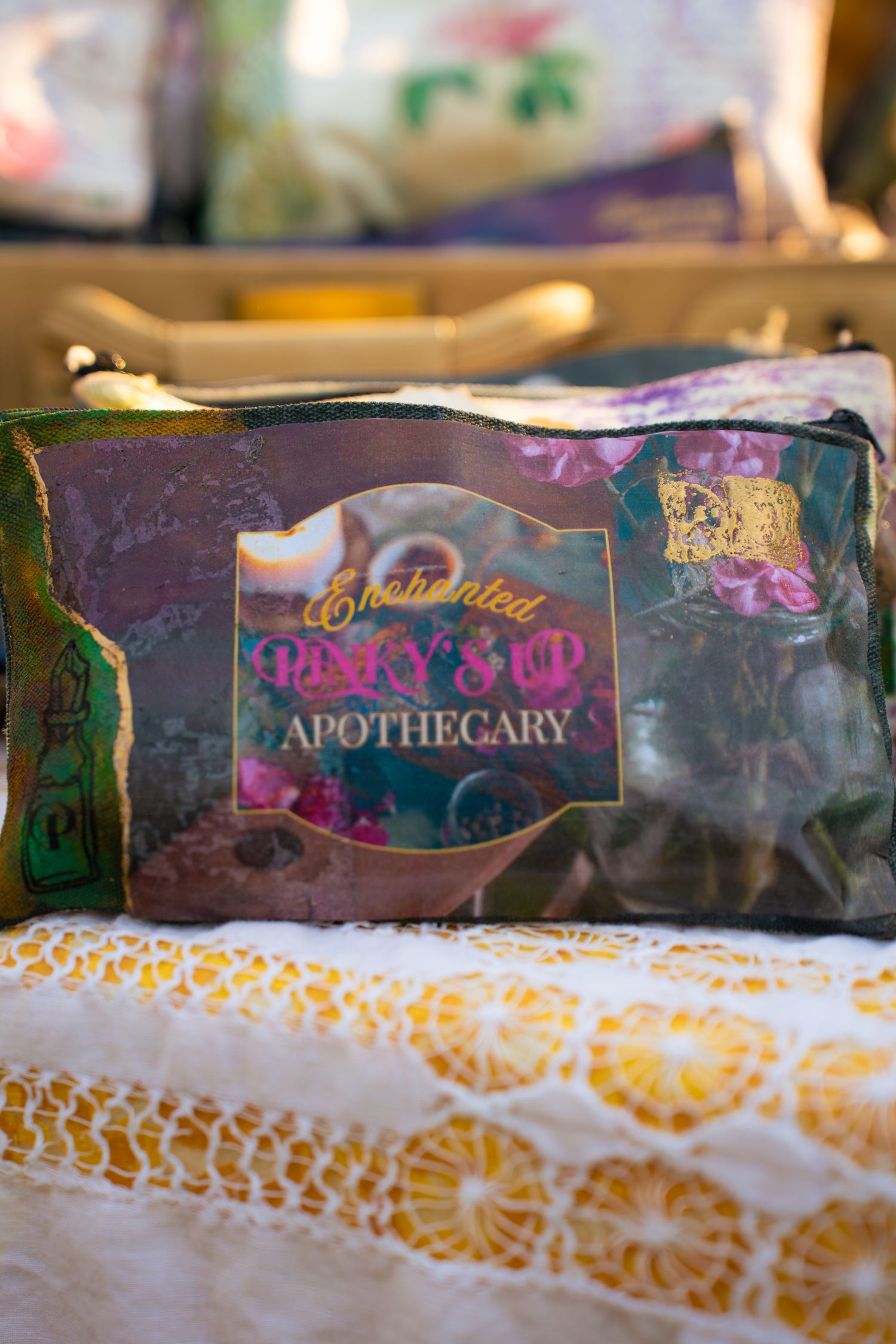 Enchanted Pixy's Sip Apothecary floral-print pouch with purple and green design resting on white lace with orange medallion pattern.
