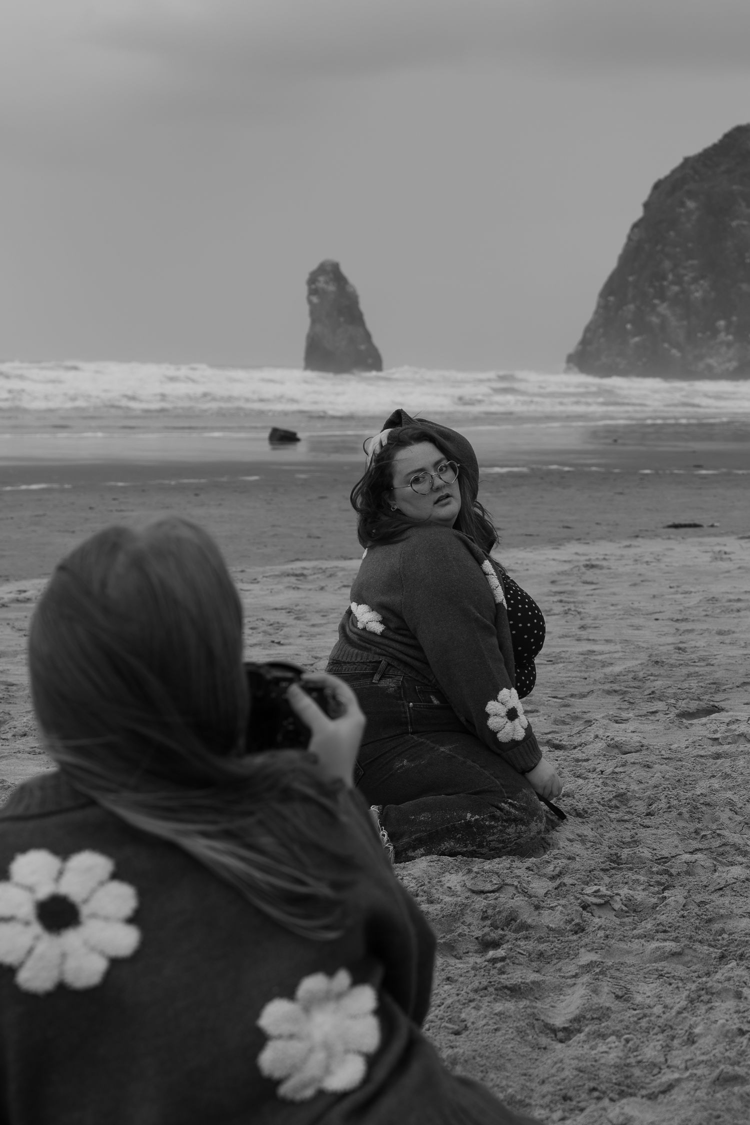 Black and white photo of woman being photographed on sandy beach with dramatic rock formations in background.