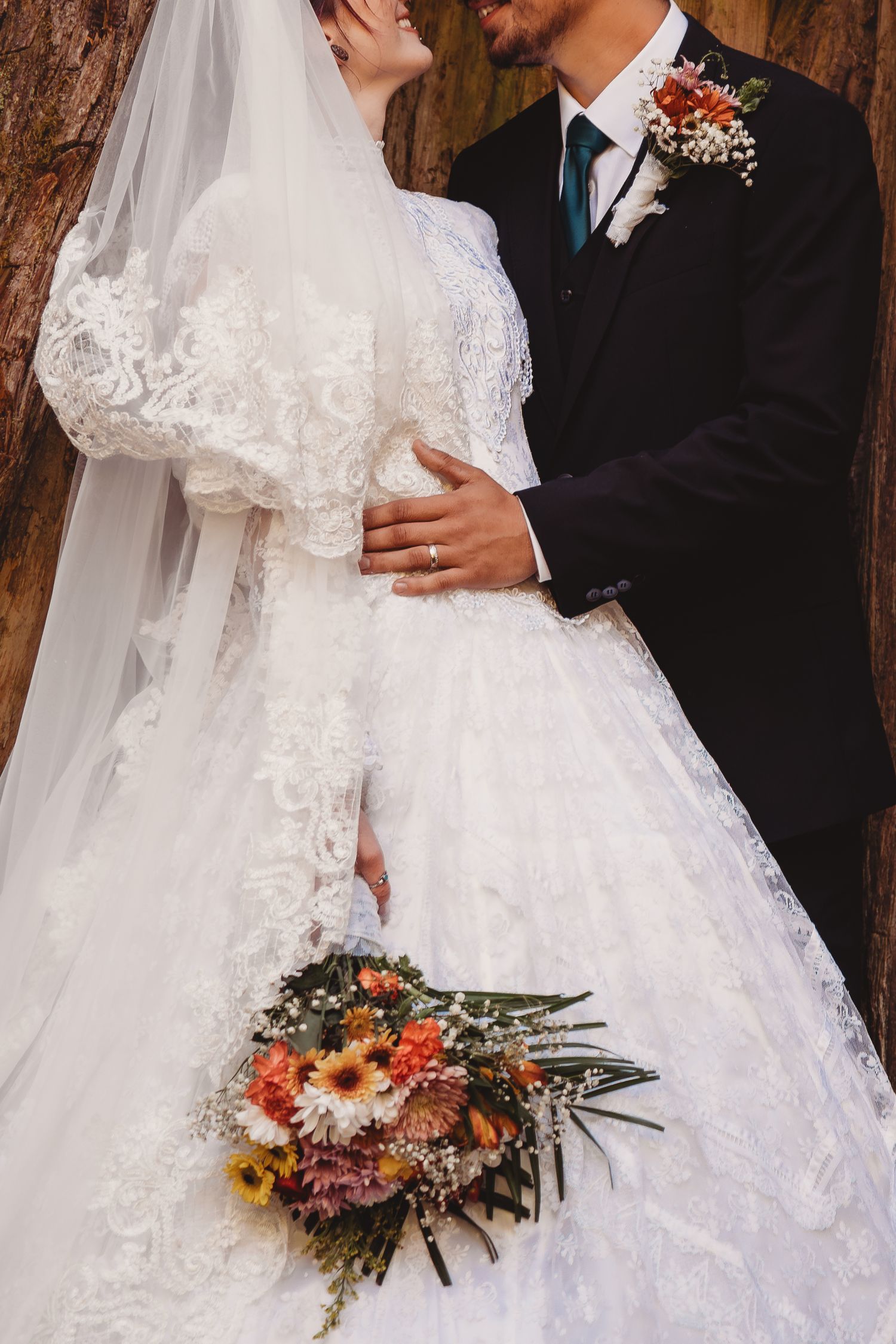Bride in lace wedding gown with veil holds colorful bouquet beside groom in black suit with boutonniere.