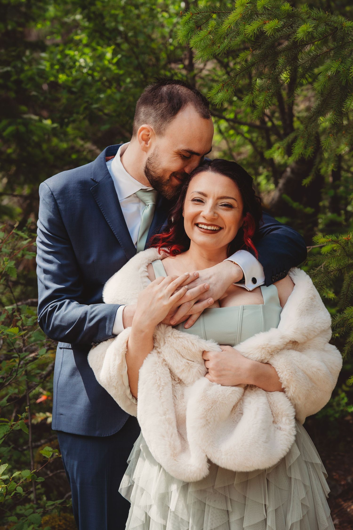 A groom in a blue suit embraces his smiling bride in a fur wrap and mint dress among lush green trees.