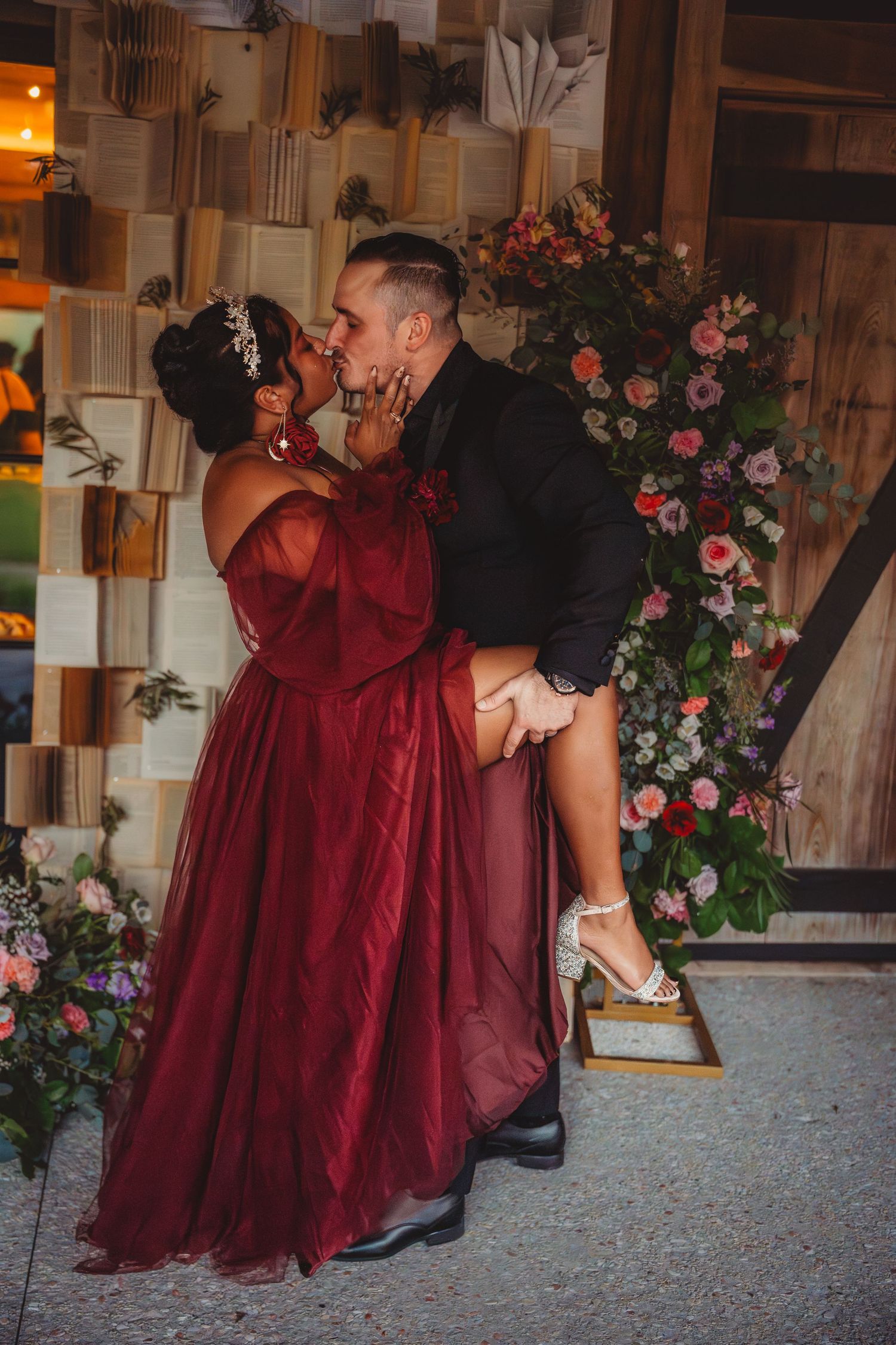 Couple sharing a romantic kiss at a wedding, woman in red gown lifted by man in black suit, floral backdrop.