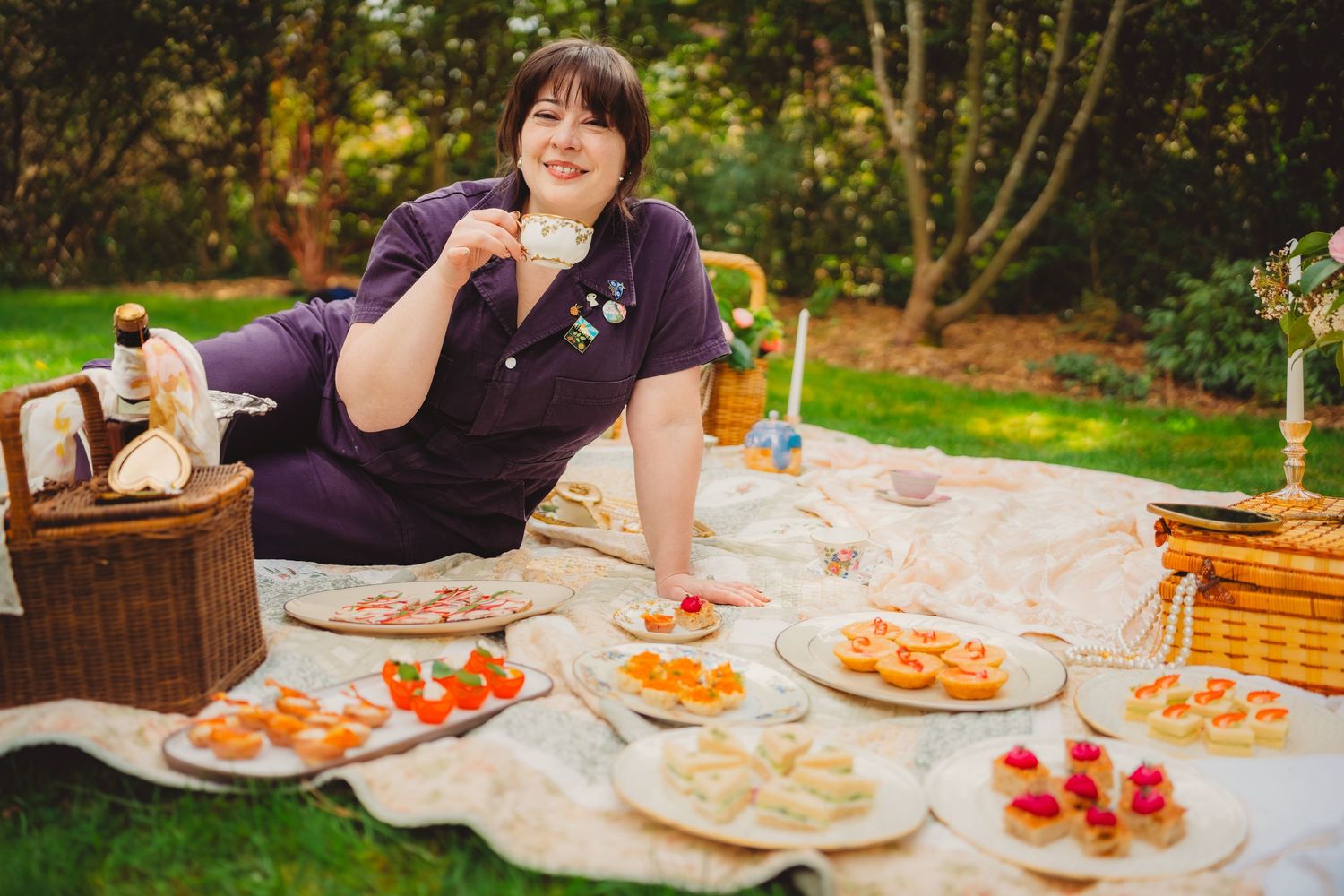 Woman enjoying a garden picnic on a blanket with assorted finger foods, sandwiches, and fruit on plates beside a wicker basket.