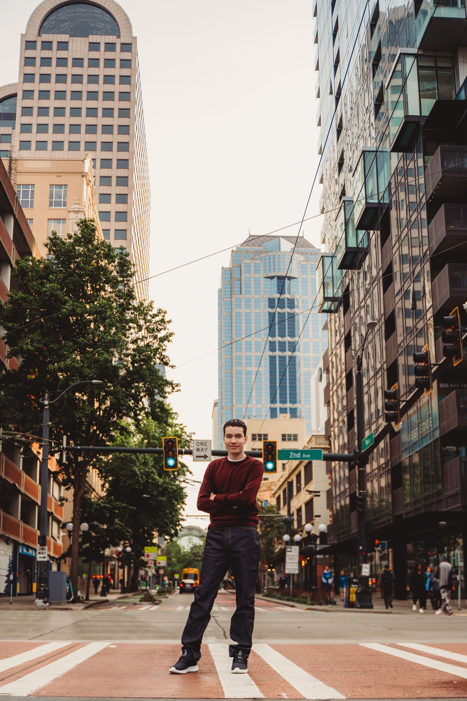 Young man standing confidently at urban crosswalk with arms crossed, surrounded by city skyscrapers and green traffic light.