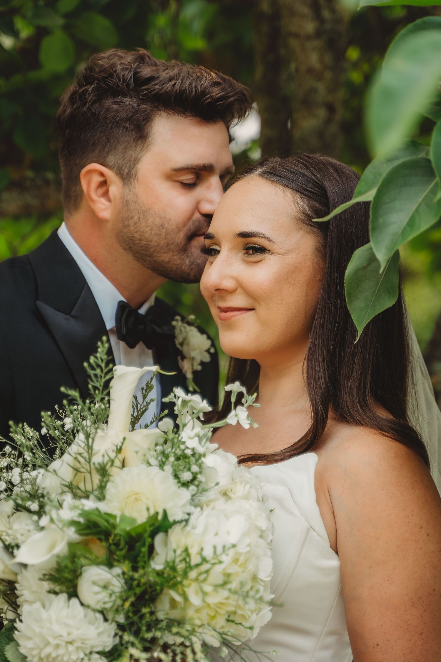 Groom in black tuxedo kissing bride on forehead while she holds white floral bouquet outdoors.Charleston South Carolina