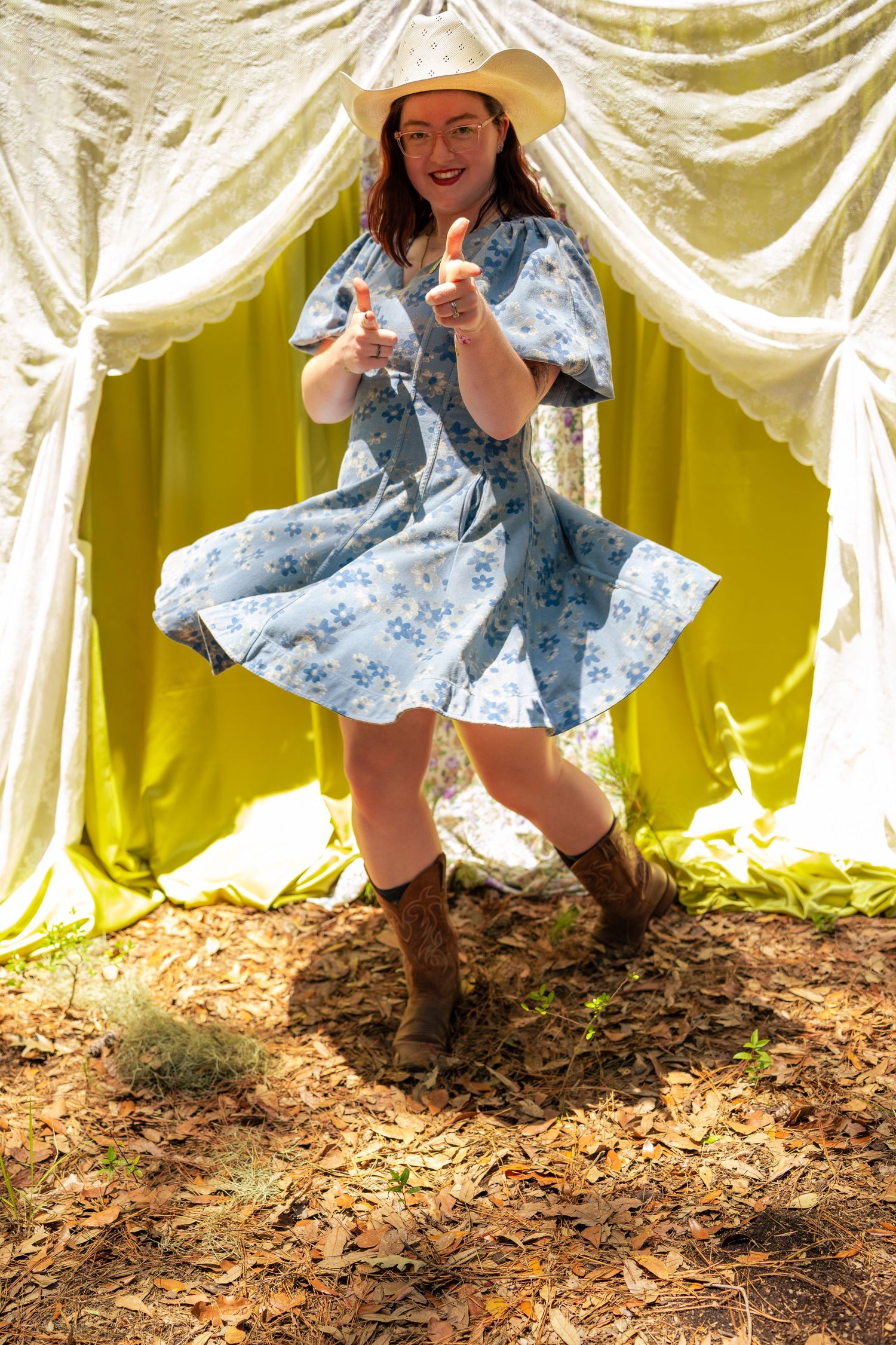 Woman in floral dress, cowboy hat, and boots dancing outdoors near colorful curtains on leaf-covered ground.