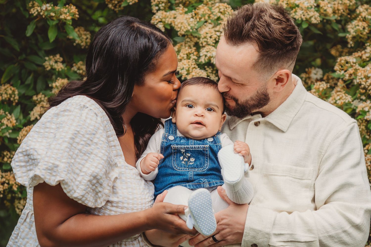A couple kisses their baby in denim overalls outdoors with blooming white flowers in the background.