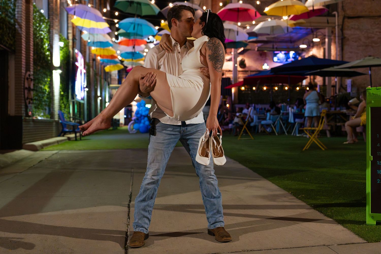 Couple kissing as a man lifts a woman in a white dress off the ground on an outdoor walkway beneath colorful hanging umbrellas at dusk.