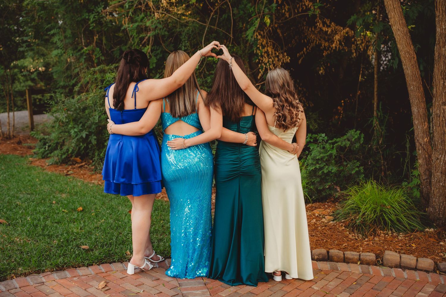 Four girls in formal dresses making a heart shape with their hands while standing together outdoors.