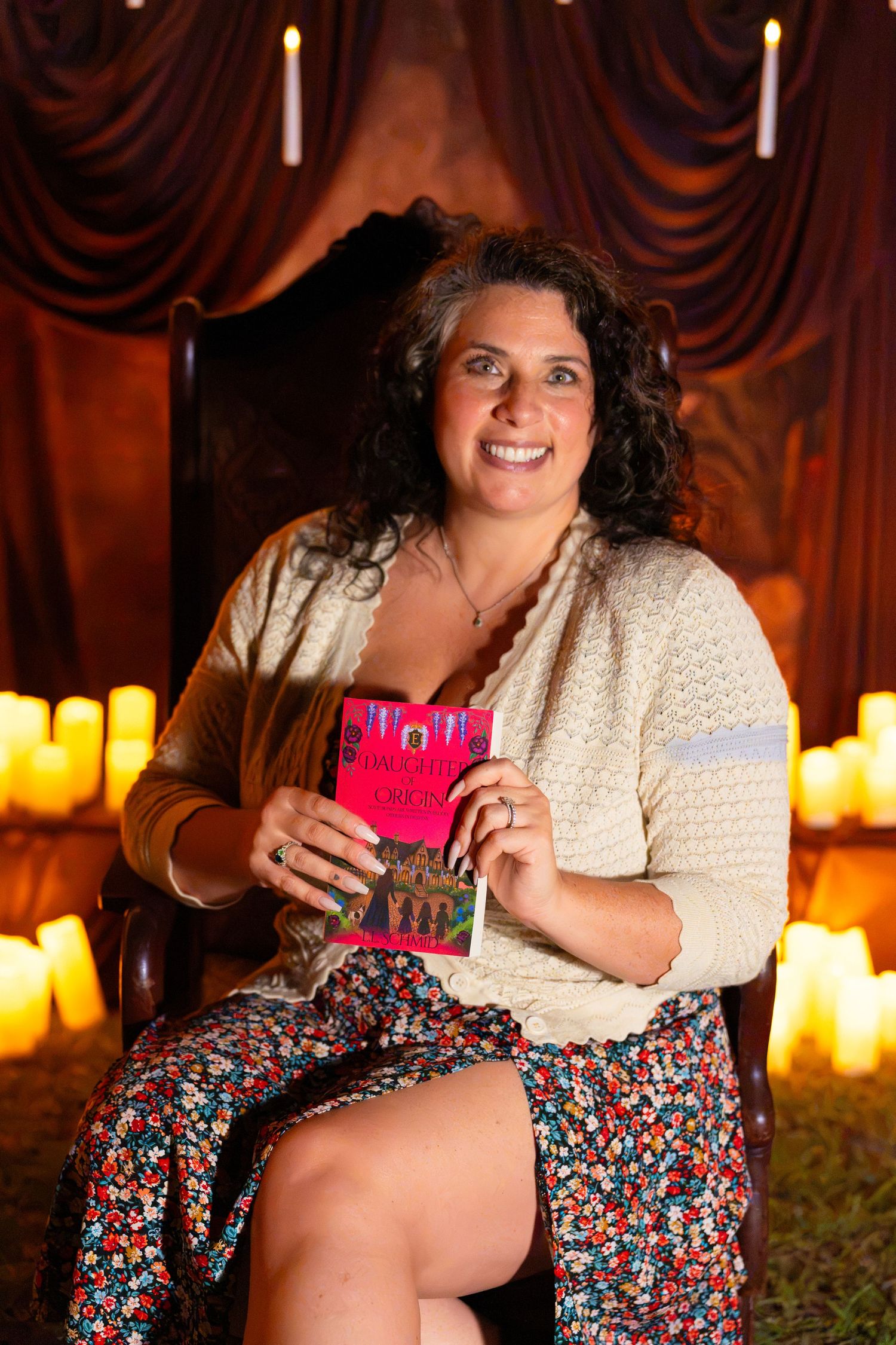 Smiling woman with curly dark hair sits in an ornate chair holding a pink book, surrounded by glowing candles and draped curtains.