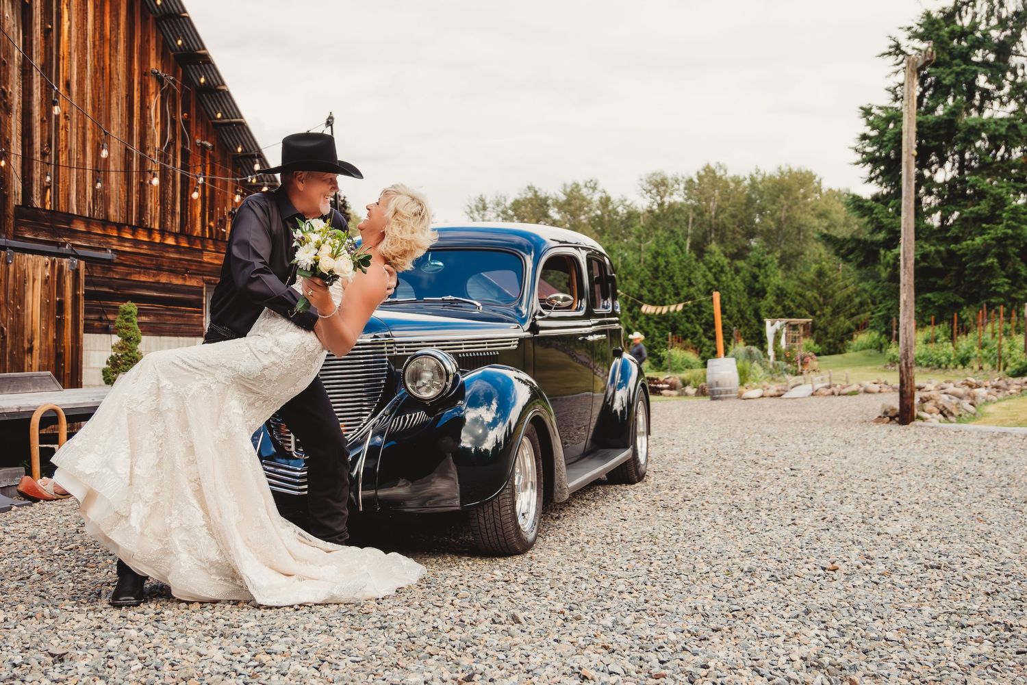 Bride and groom share a romantic dip kiss beside a classic black vintage car on a gravel drive at a rustic barn wedding.