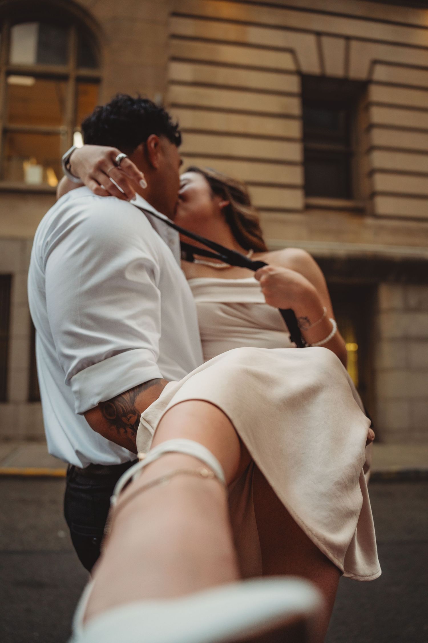 A couple shares a romantic kiss outdoors, with the man lifting the woman in a white dress while she pulls his tie.