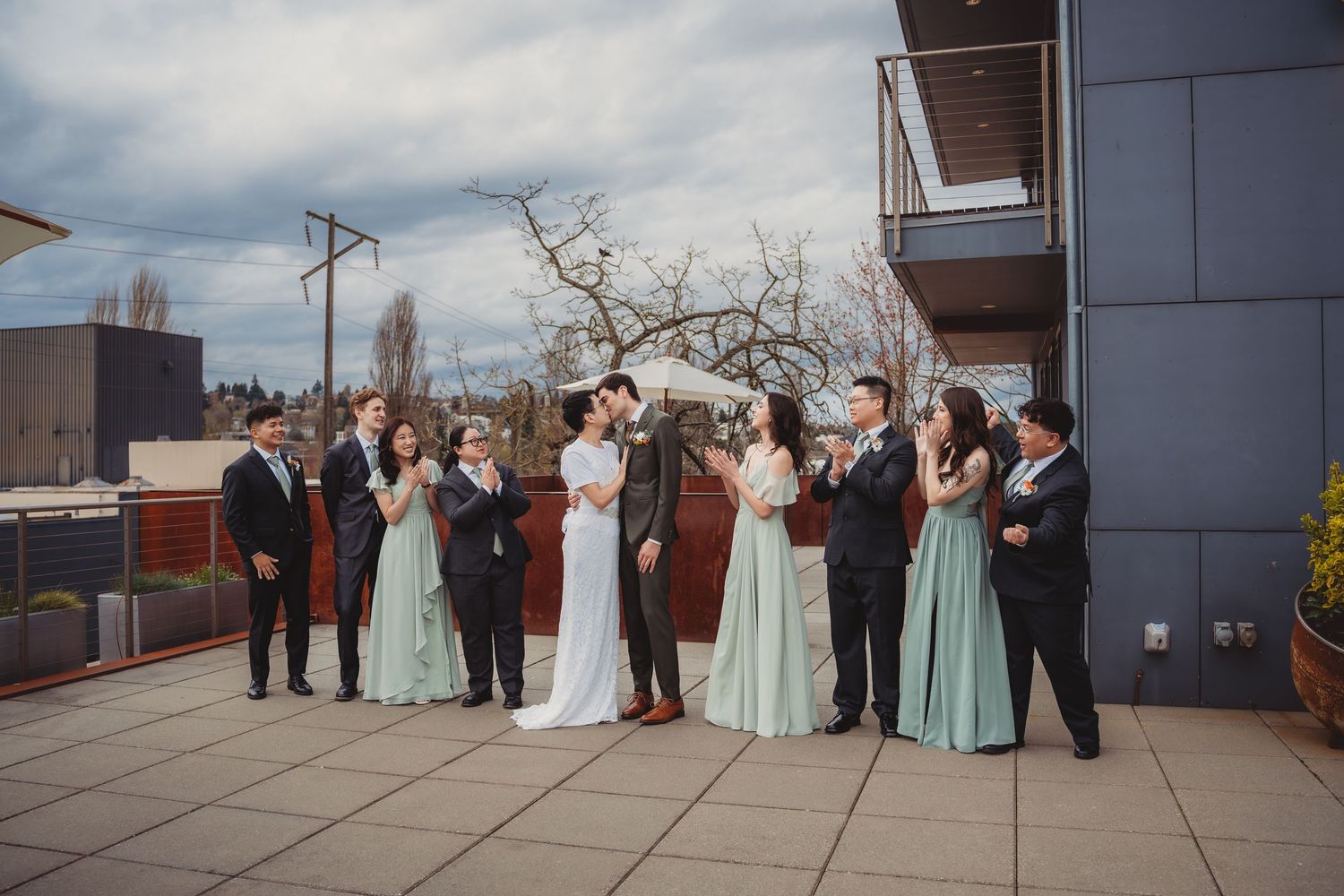 Wedding party celebrating on a rooftop terrace, bride and groom kissing while guests in teal gowns and suits applaud.