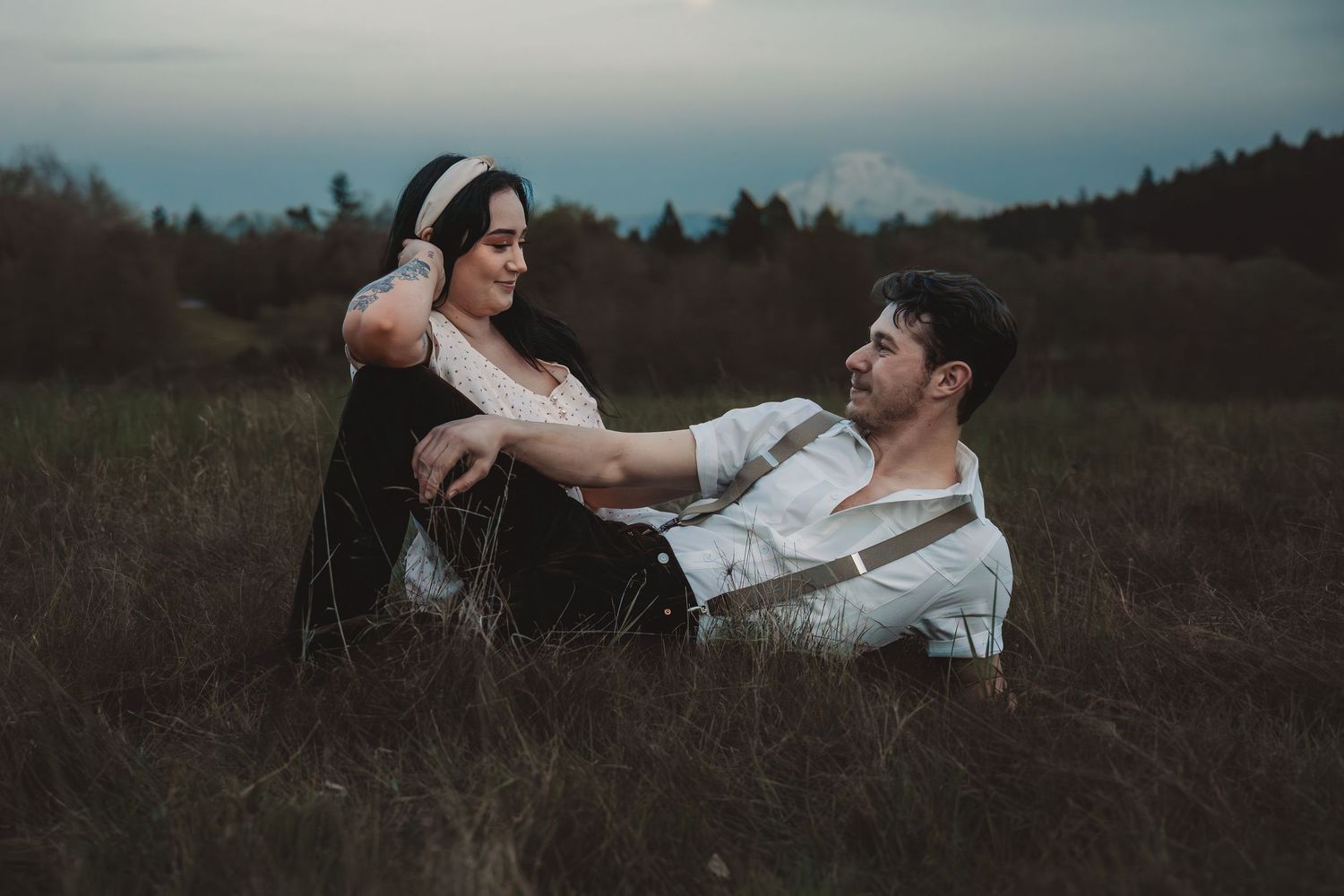 Couple sitting intimately in a grassy field at dusk, sharing a romantic moment in a moody outdoor setting.