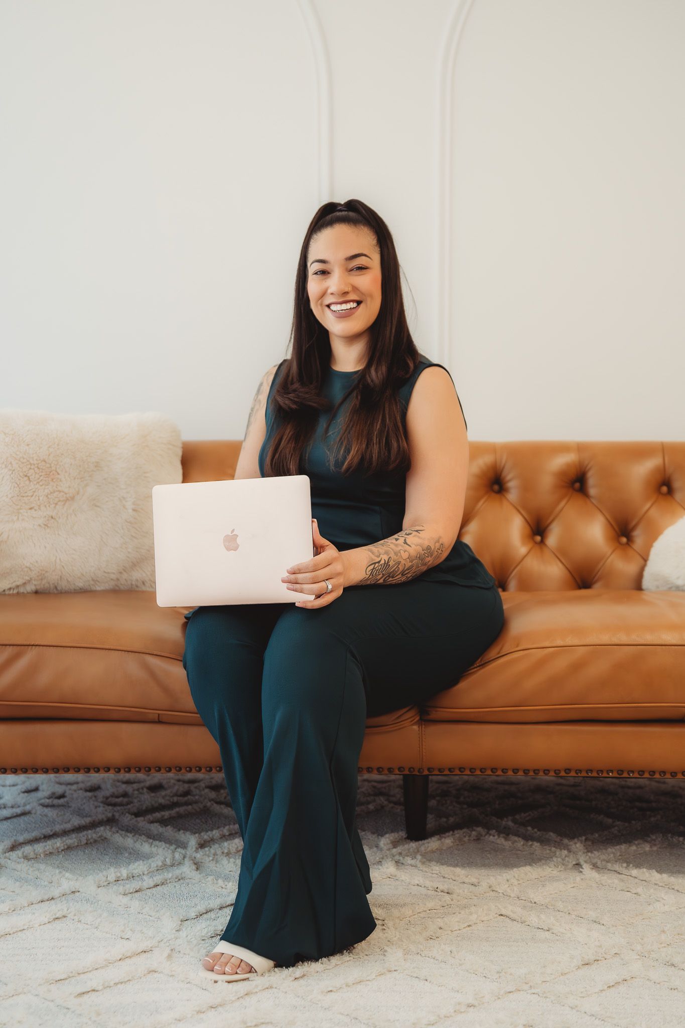 Smiling woman in dark green jumpsuit holding MacBook laptop while sitting on brown leather tufted sofa.