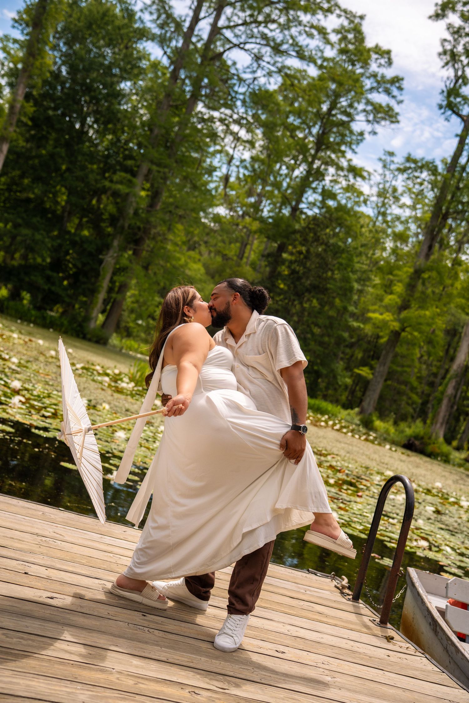Couple sharing a romantic kiss on a wooden dock surrounded by a lily pad pond and lush green trees.