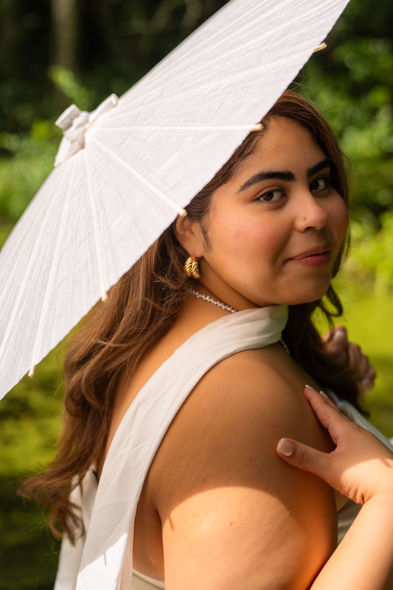 Young woman in white dress holding a white parasol umbrella outdoors, with lush green foliage background.