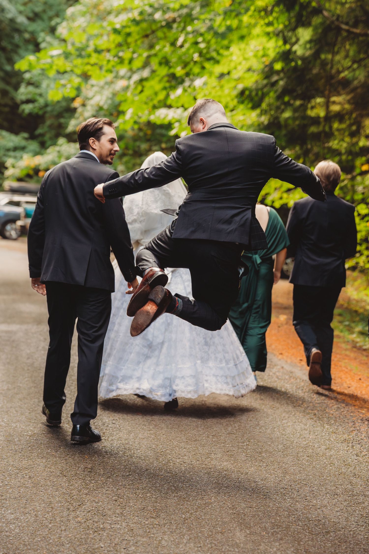 Groom jumping joyfully with wedding party walking on a tree-lined path in formal attire.