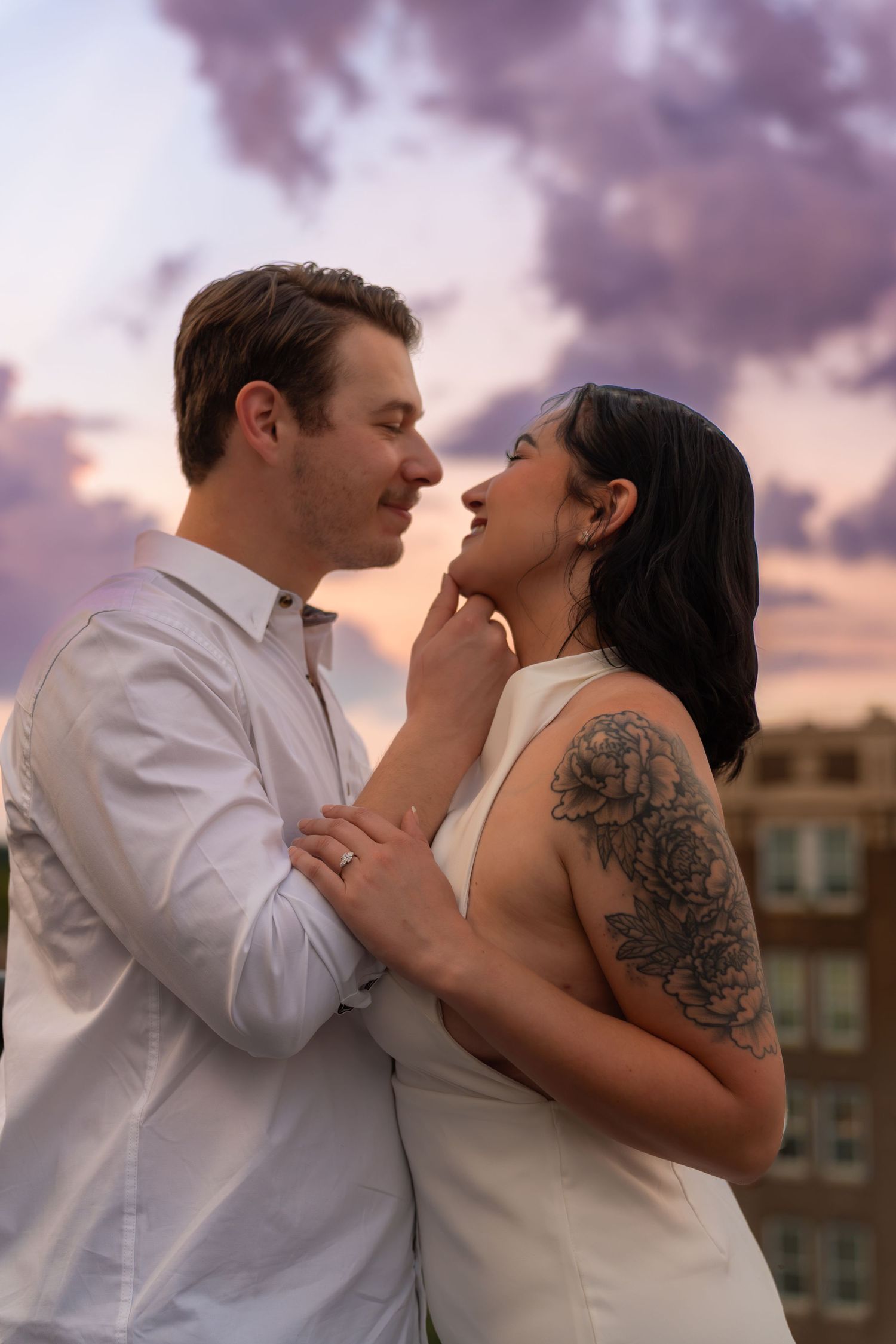 Couple embracing at sunset on a rooftop, the woman with a floral shoulder tattoo smiling as the man gently touches her chin.