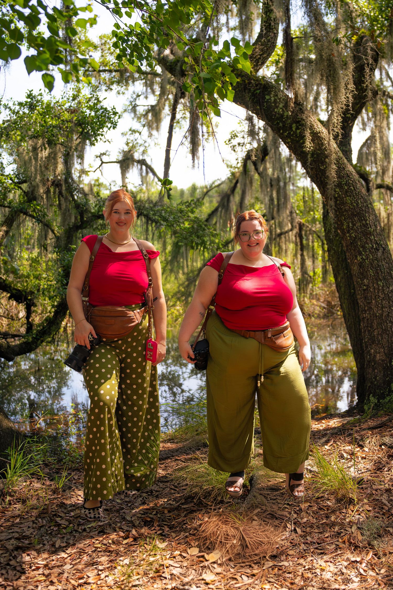 Two women in matching red tops and green pants pose in a mossy Southern swamp forest setting.