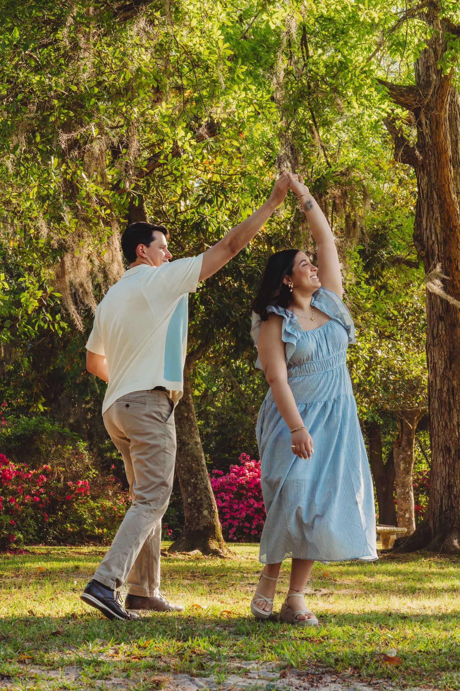 Couple dancing outdoors in a lush green park, man spinning woman in a blue dress under sunlit trees.
