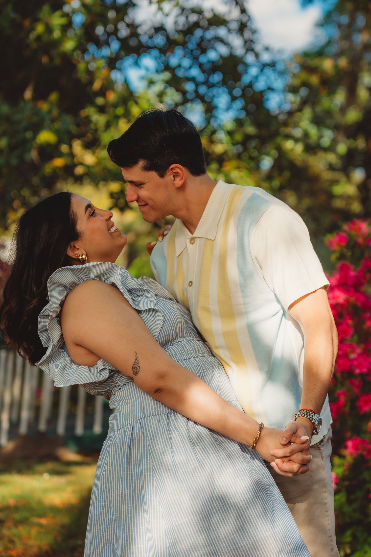 A couple shares a romantic moment in a sunny garden, smiling at each other surrounded by colorful flowers and lush greenery.