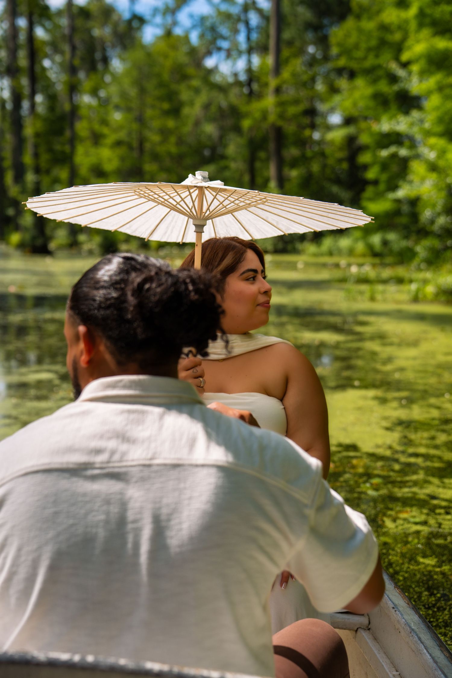 Couple on a boat in a lush green swamp, woman in white dress holding a parasol umbrella on a sunny day.