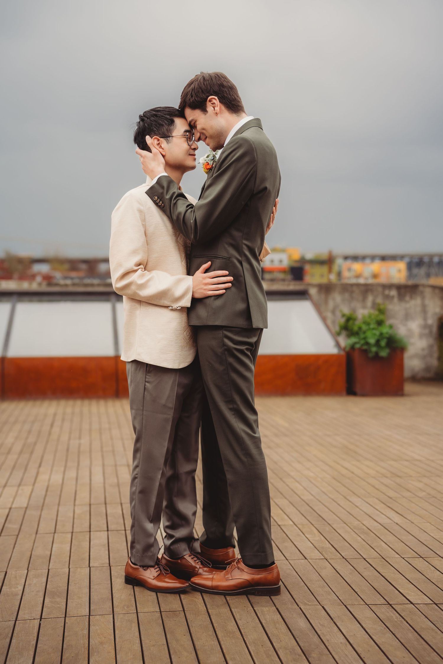 Two grooms share an intimate moment on rooftop, one in cream jacket and one in olive suit, foreheads touching tenderly.