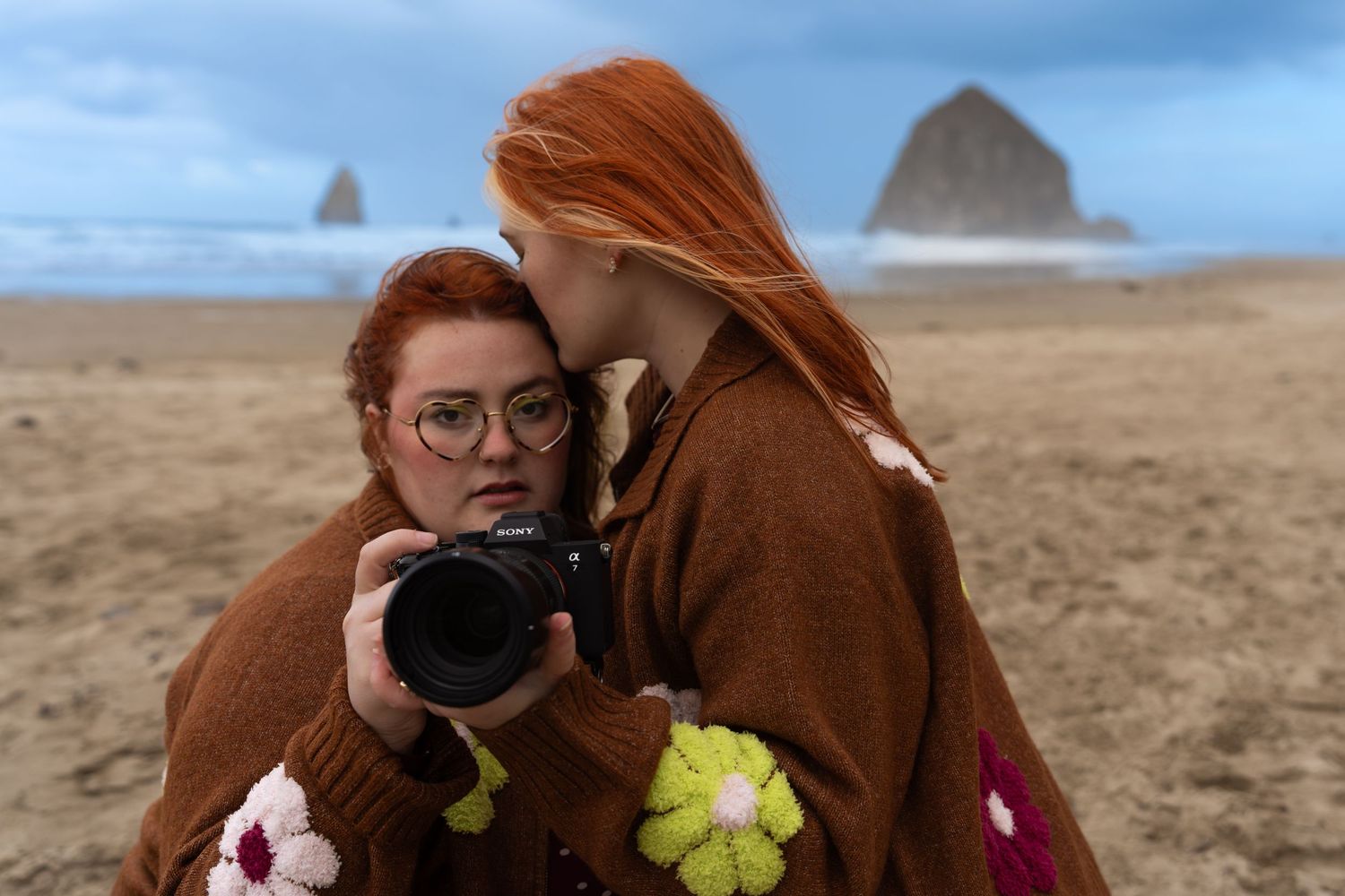Two redhead women in brown sweaters examine a camera on a sandy beach with dramatic rock formations in the background.