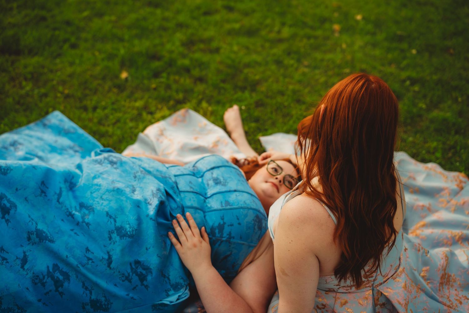 Two redhead women lying on a blue blanket in the grass, sharing a tender moment outdoors.