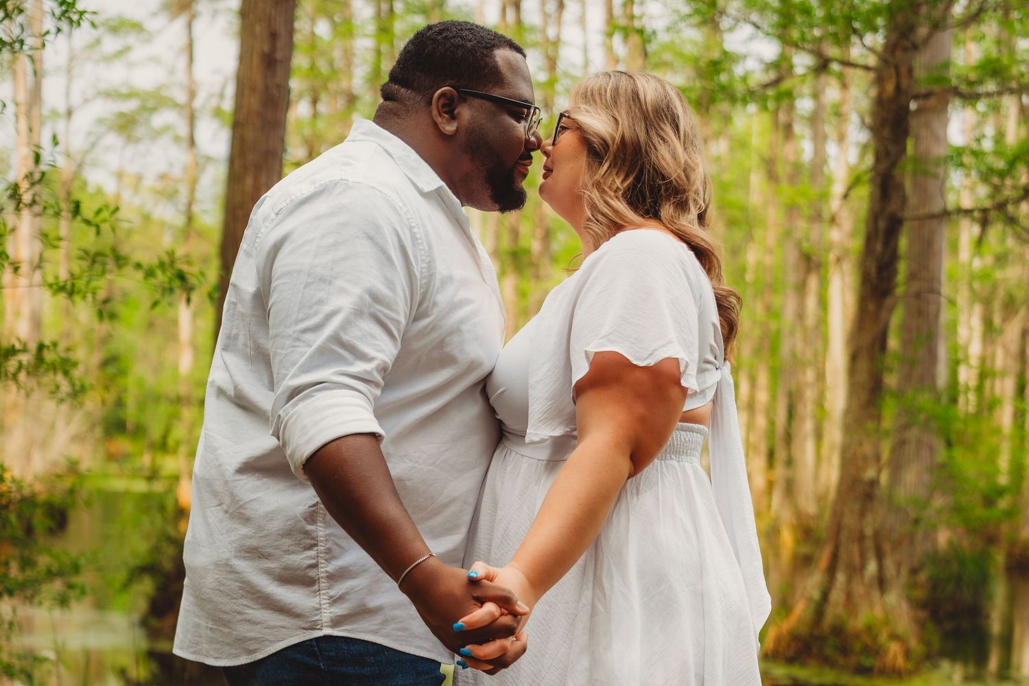 Interracial couple sharing a kiss while holding hands outdoors in a lush green forest setting.