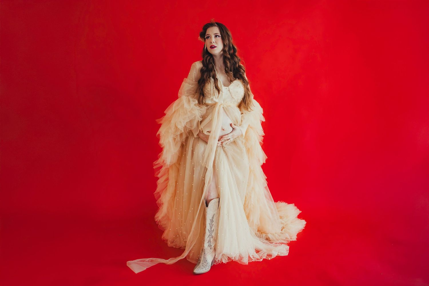 Woman in elegant cream ruffled gown posing against vibrant red background in a dramatic fashion portrait.