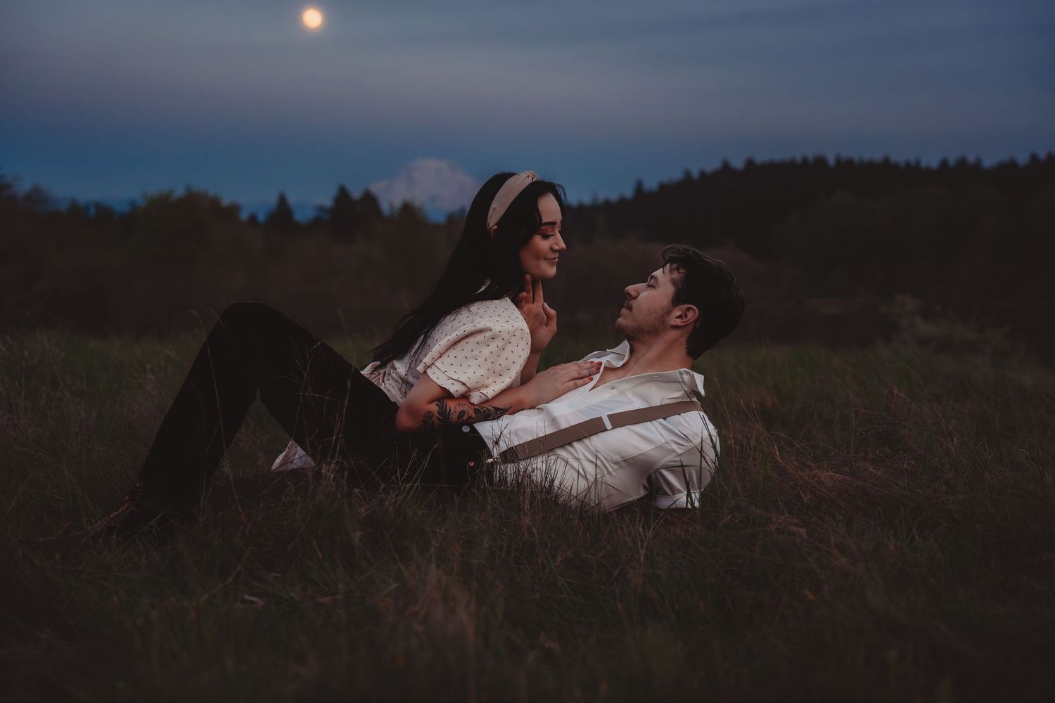 Romantic couple sitting together in a moonlit field at dusk, sharing a tender moment outdoors.