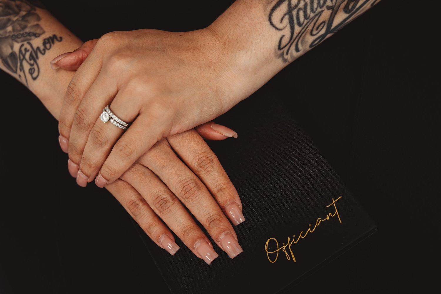 Close-up of tattooed hands with diamond ring resting on black officiant book against dark background.