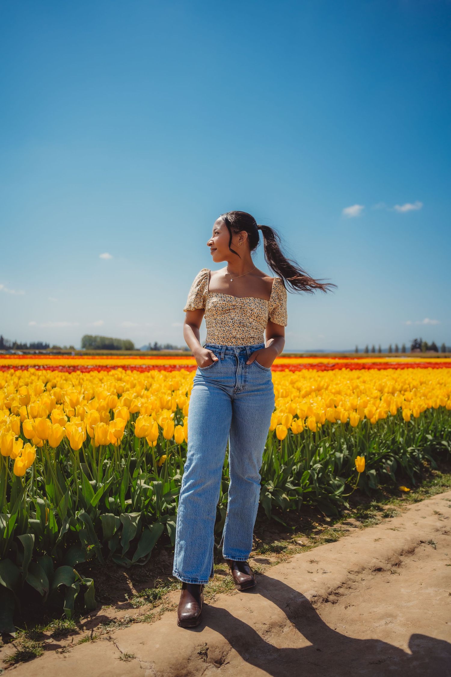 Woman in floral top and jeans standing in vibrant yellow and red tulip field under clear blue sky.