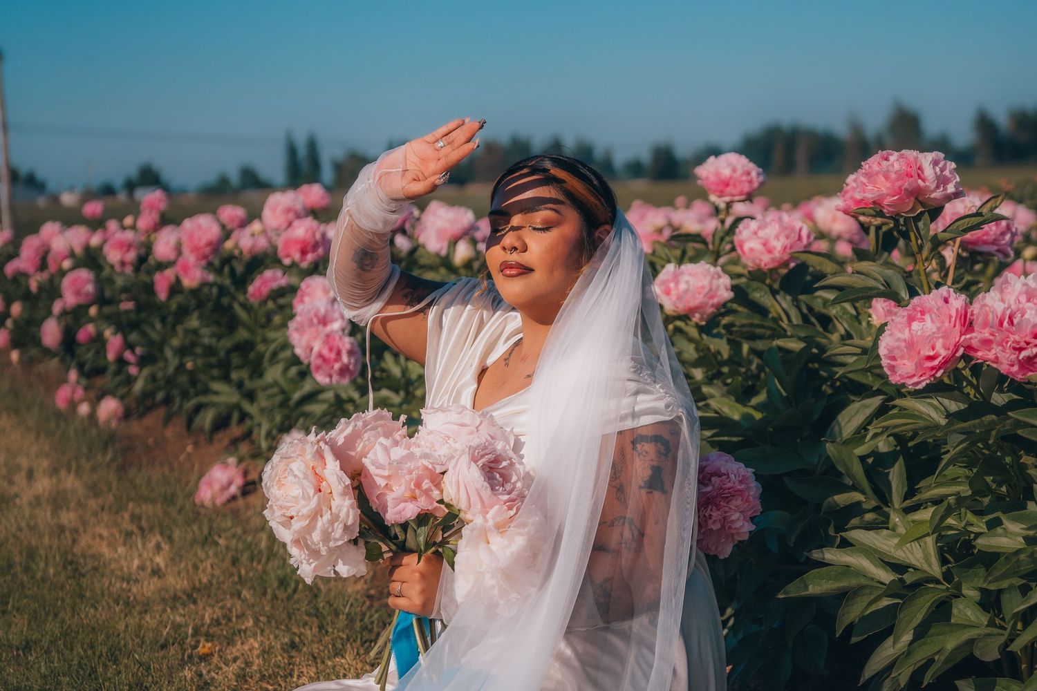 Bride in white gown and veil holds pink peonies bouquet while shielding eyes from sun in blooming flower field.