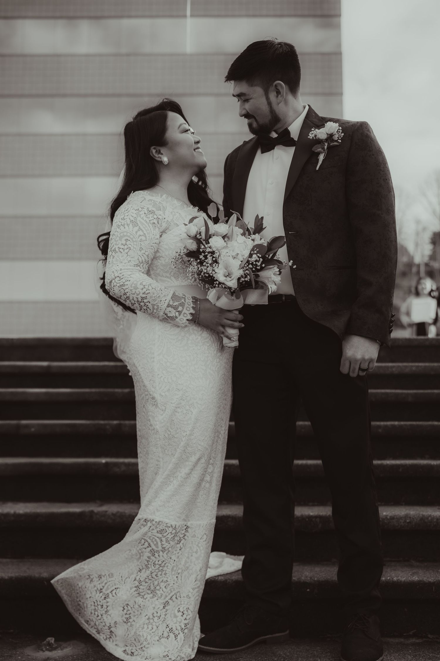 Black and white photo of bride in lace gown and groom in tuxedo sharing a romantic moment on steps.