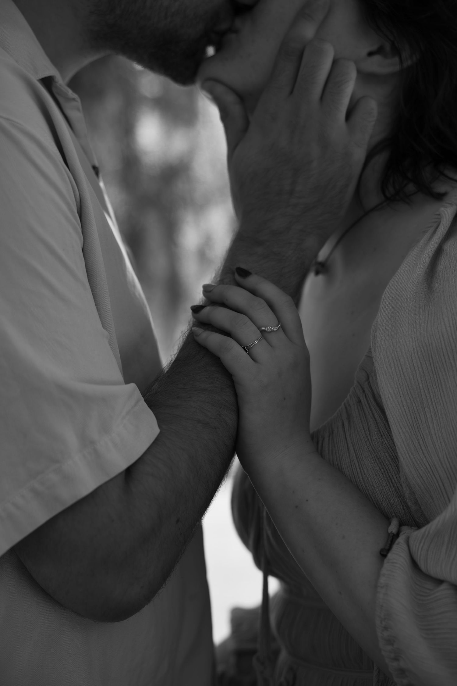 Black and white photo of a couple sharing a tender kiss, hands intertwined with an engagement ring visible.