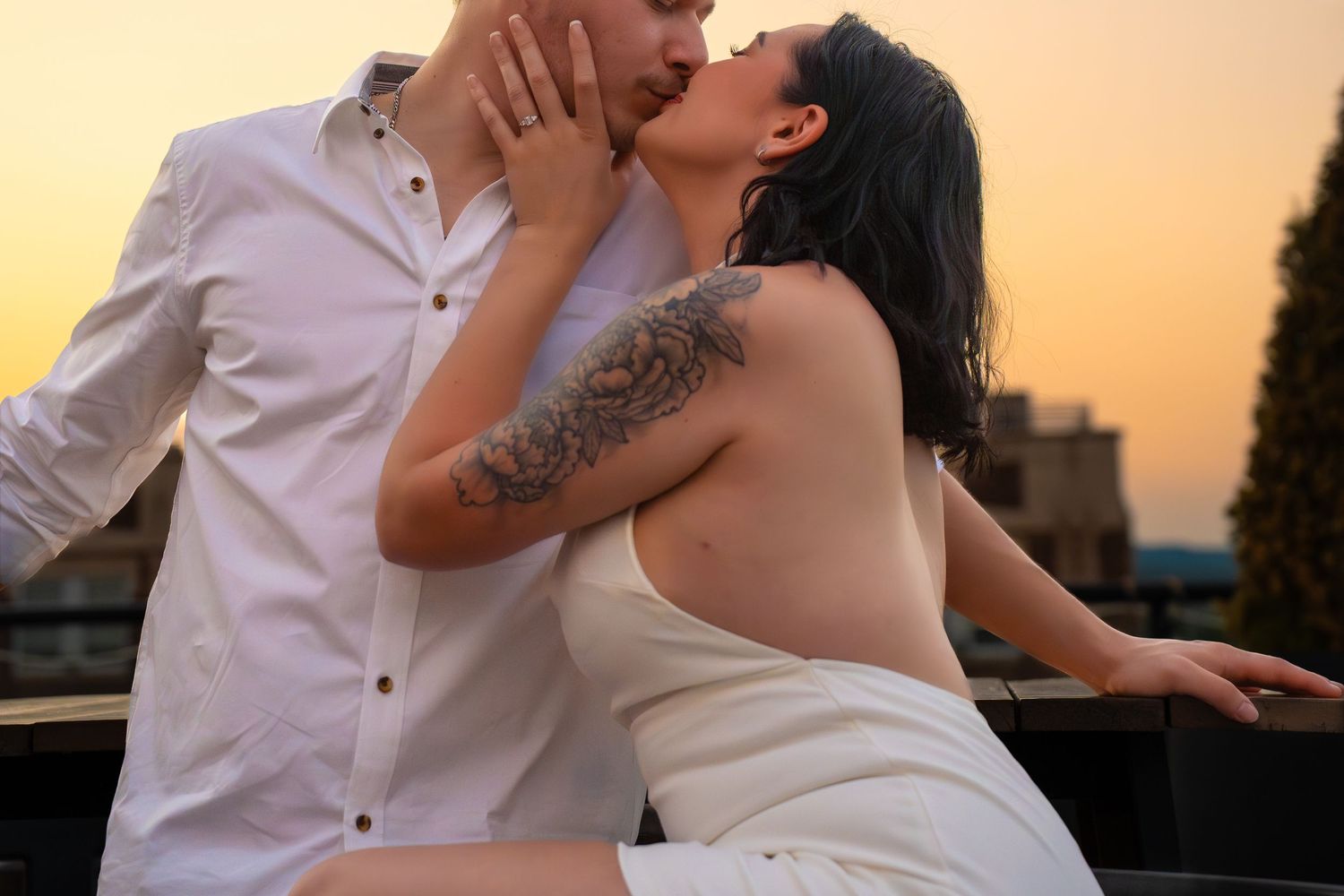 Romantic couple kissing on a rooftop at sunset, the woman in a white dress with a shoulder tattoo and the man in a white shirt.