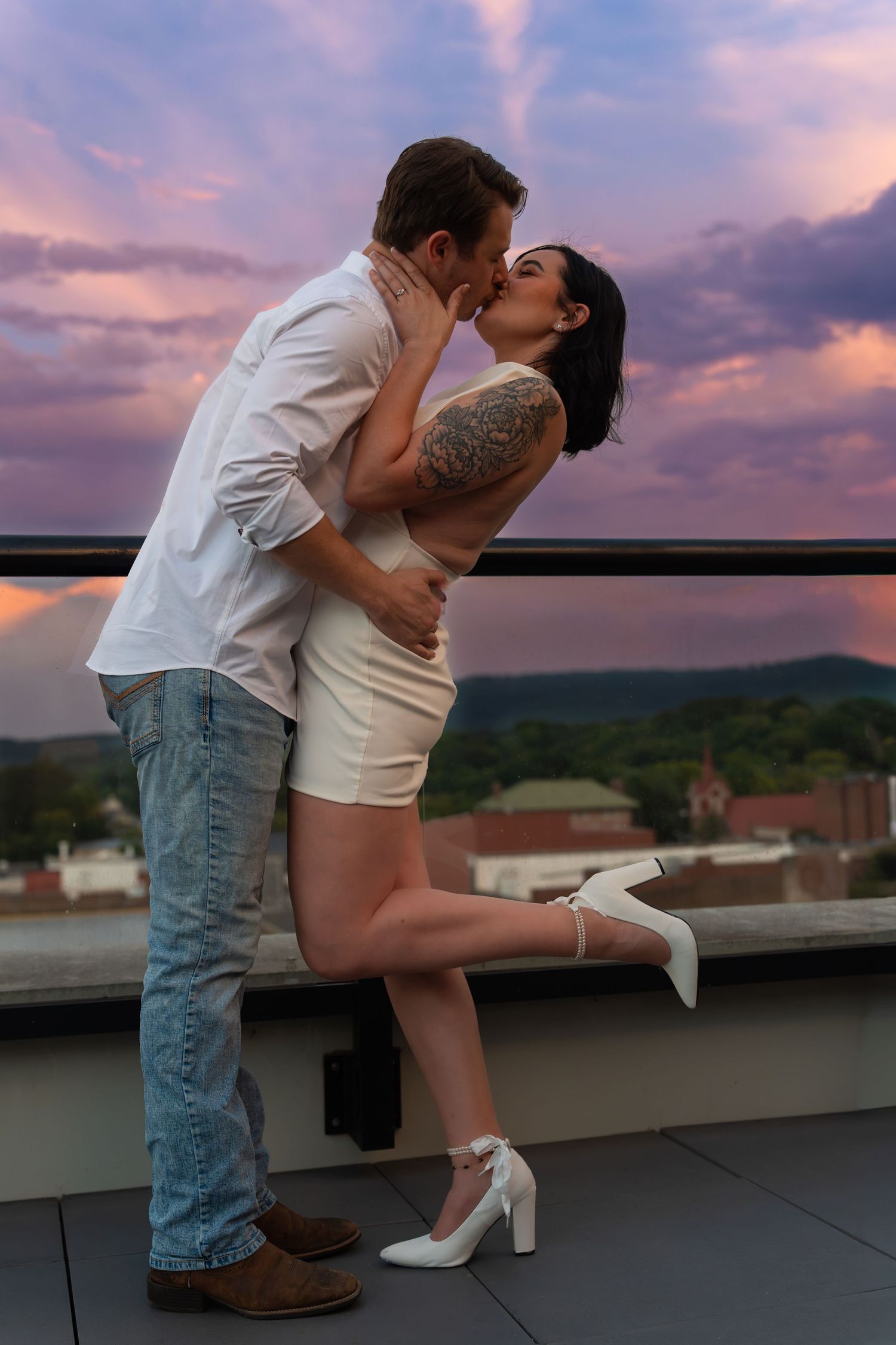 Couple kissing on a rooftop terrace at sunset, she in a white dress and heels with an arm tattoo, with purple clouds and town view.