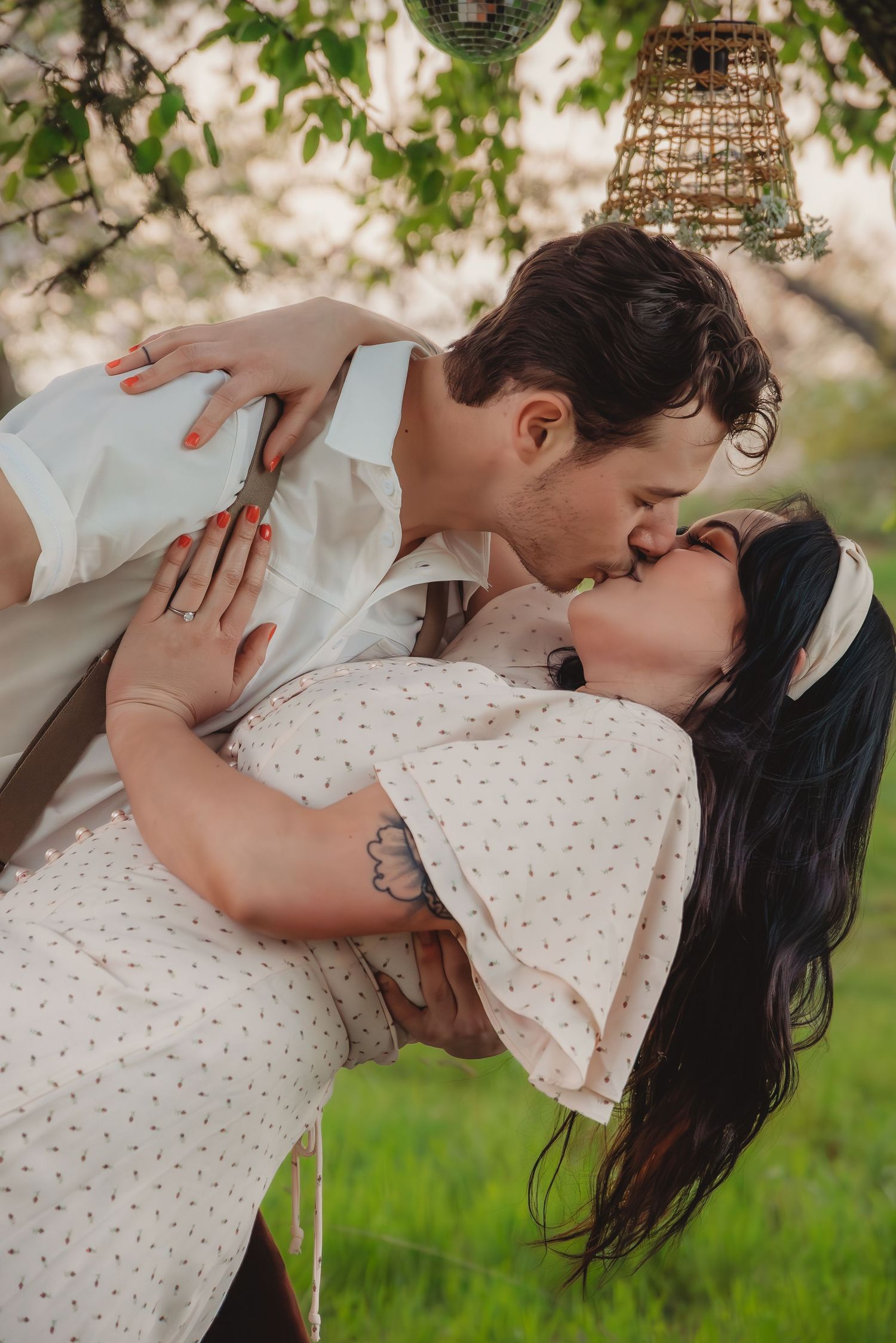 Couple sharing a passionate kiss outdoors, woman in polka dot dress dipped back under blooming tree.